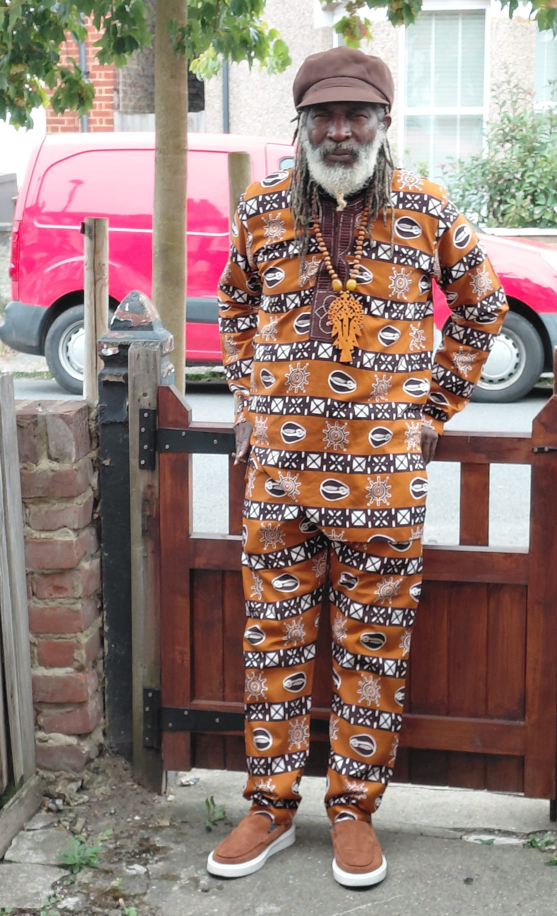 Fyah Mawi wearing a patterned suit, standing beside a garden gate.