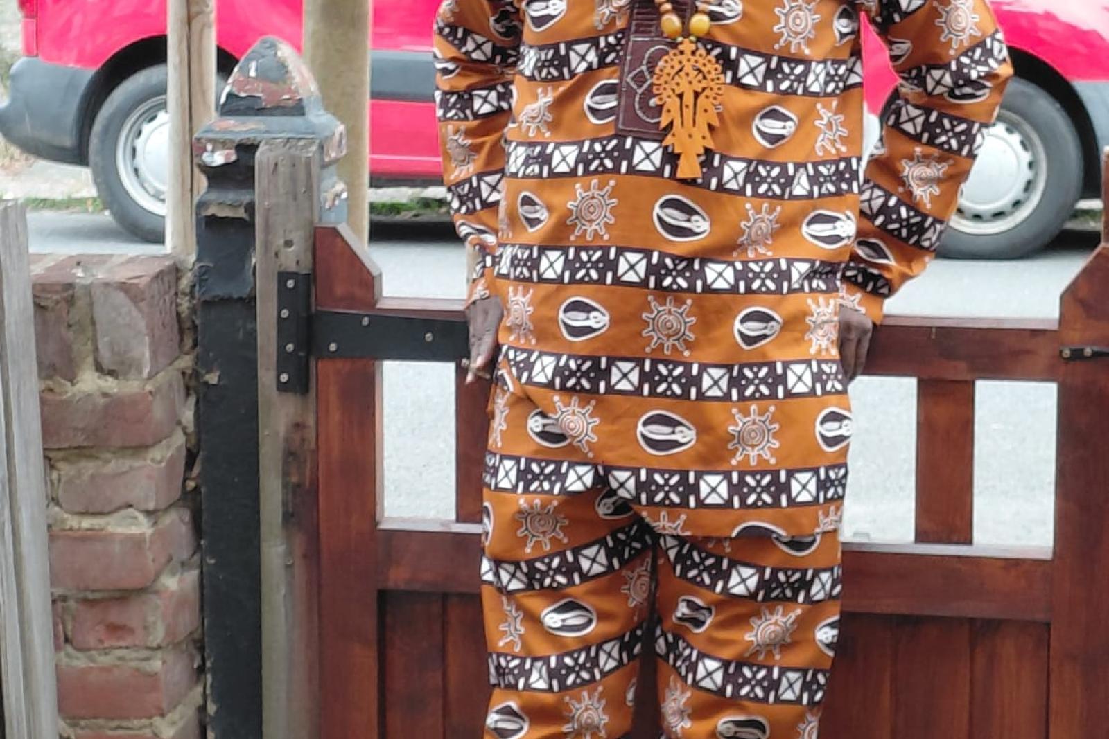 Fyah Mawi wearing a patterned suit, standing beside a garden gate.