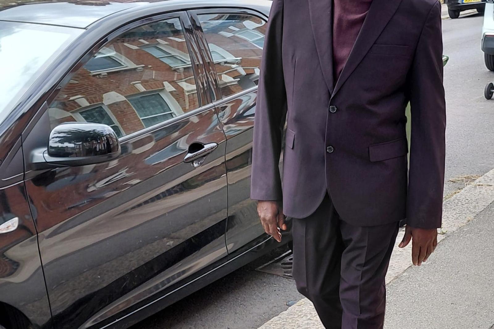 Fyah Mawi walking beside a parked car in a dark suit.