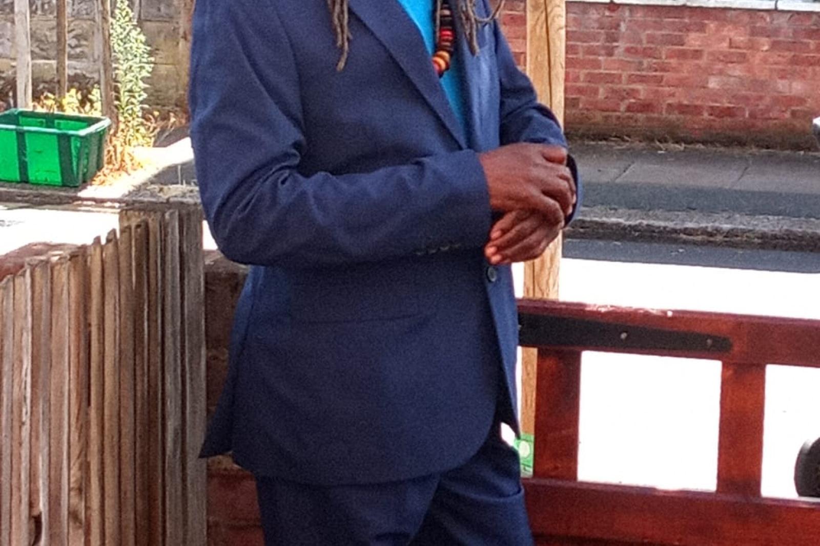 Fyah Mawi in a blue suit and wide-brimmed hat by a gate.
