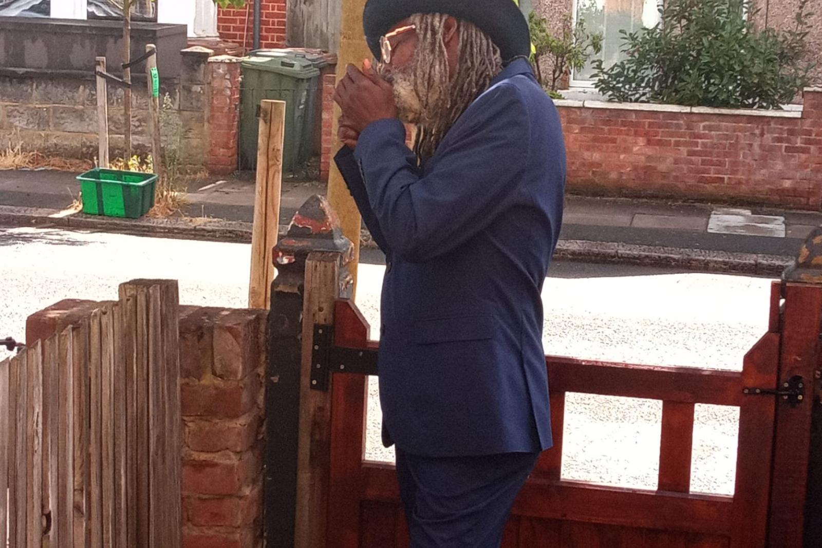 Fyah Mawi in a blue suit leaning beside a garden gate.
